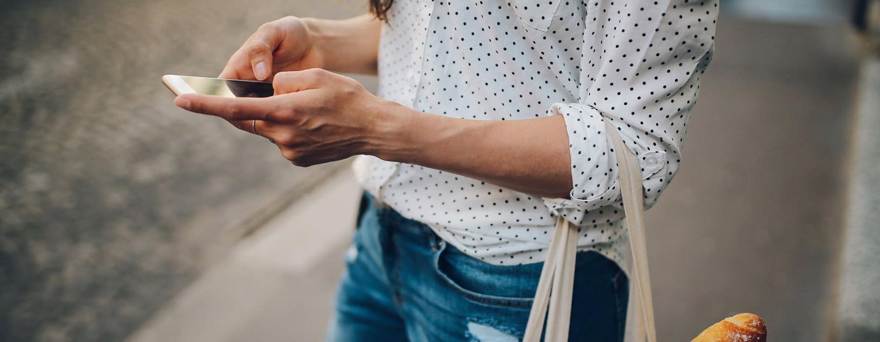 woman walks down the street and texts with a bag of groceries on her arm